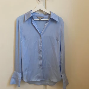 Ljusblå skjorta - Snygg ljusblå skjorta med klassisk krage och knappar framtill. Materialet är slätt och har en lätt glans, vilket gör att den känns extra fräsch. Perfekt för dig som gillar stilrena och enkla plagg. Säljer då den är för liten för mig.