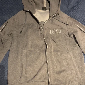 Grå hoodie från Hugo Boss, M - Säljer en grå hoodie från Hugo Boss i storlek M. Tröjan har dragkedja, huva med snörning och logga på bröstet. Regular fit och praktiska fickor framtill. Perfekt för en chill och stilren look. PASSAR OM DU HAR STORLEK S