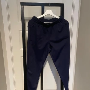 Mörkblå mjukisbyxor från Under Armour - Mörkblå mjukisbyxor från Under Armour med elastisk midja och sidofickor. Byxorna har en avslappnad loose fit och är tillverkade i mjukt syntetmaterial, perfekt för chill eller träning. Under Armour-logga på benet för en sportig vibe.