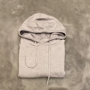 Ljusgrå hoodie med luva och snörning - En stilren ljusgrå hoodie med klassisk huva och snörning. Tröjan har långa ärmar och ribbade muddar vid ärmslut och nederkant. Perfekt för chill dagar eller när du vill ha en enkel och clean look.