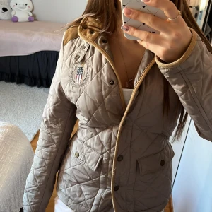 Beige Lexington jacka - Snygg beige jacka från Lexington med bruna axelpartier, broderad logga på bröstet och detaljer i mockaimitation. Jackan har knappar framtill, två stora fickor och en klassisk krage. 