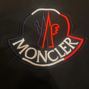 Svart Moncler sweatshirt med stor logga - Svart sweatshirt från Moncler med stor broderad logga i vitt, rött och blått på bröstet. Tröjan har rund halsringning och långa ärmar. Perfekt för dig som gillar streetwear och vill sticka ut med en ikonisk design.