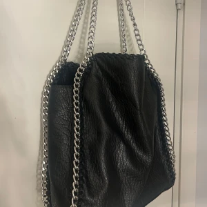 Svart axelväska med kedjor från Tiamo - Cool svart axelväska från Tiamo i skinnimitation med grova silverfärgade kedjor som handtag och detaljer längs kanterna. Väskan har ett rymligt huvudfack, innerficka med dragkedja och snyggt texturerad yta. Perfekt för dig som gillar edgy stil och vill ha plats för allt.