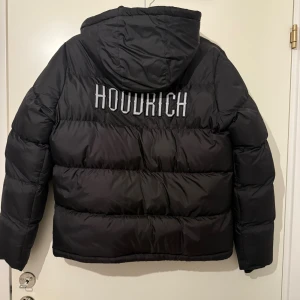 Svart pufferjacka från Hoodrich XL(ungdom) - Svart pufferjacka från Hoodrich med stor broderad logga på ryggen och mindre logga på bröstet. Jackan har huva, dragkedja framtill och är vadderad för extra värme. Inköpt på jdsport förra vintern!