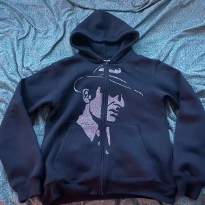 Svart hoodie med tryck från Costino - Svart hoodie från Costino Stockholm med dragkedja och huva. Framsidan har ett stort tryck av en person med hatt i vitt. Mjuk insida och kängurufickor framtill. Perfekt för dig som gillar streetstyle och vill sticka ut.