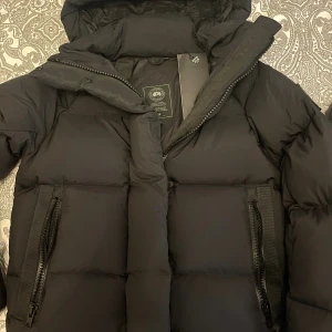 Svart dunjacka från Canada Goose - Säljer en helt ny svart dunjacka från Canada Goose med huva och diskret logga på ärmen. Jackan har två stora fickor med dragkedja framtill och en reflekterande detalj på huvan. Perfekt för kalla vinterdagar och riktigt snygg passform, lite kortare modell.