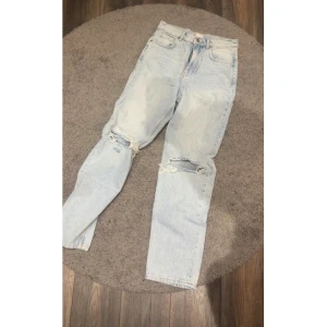 Ljusblå slitna raka jeans - Säljer ett par ljusblå jeans med raka ben och slitna detaljer vid knäna. Jeansen har klassisk femficksdesign och är tillverkade i denim. Perfekta för en avslappnad och trendig look.