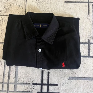 Svart skjorta från Ralph Lauren - Klassisk svart skjorta från Ralph Lauren med röd broderad logga på bröstet. Skjortan har knappar framtill och är tillverkad i mjuk bomull. Perfekt för dig som gillar stilrena och tidlösa plagg.