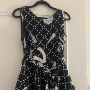 Svart byxdress  med läppstiftsmönster - Säljer en svart byxdress med coolt vitt och rött mönster av läppstift och grafiska former. Den har ärmlös design, rund hals och volangdetaljer i kjolen. Materialet känns lätt och flowy, perfekt för dig som gillar att sticka ut med din stil. Byxdress med shorts nere 