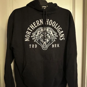 Mörk grå hoodie Northern Hooligans - Mörk grå hoodie från Northern Hooligans med stor vit logga och tryck av en rytande panter på bröstet. Klassisk känguruficka framtill och snörning i huvan.