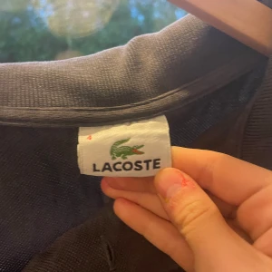 Marinblå pikétröja från Lacoste - Klassisk marinblå pikétröja från Lacoste med korta ärmar och den ikoniska gröna krokodilen broderad på bröstet. Tröjan har krage och knappar upptill. Perfekt för en clean och sportig stil.