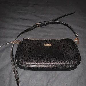 Svart axelväska från Guess - Snygg svart axelväska från Guess med guldfärgade detaljer och justerbar rem. Väskan har strukturmönstrad yta, dragkedja upptill och ett ljust innerfoder med Guess-logga. Perfekt storlek för det viktigaste och stilren look.