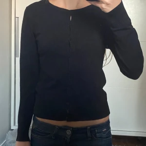 Svart kofta med knappar - En stilren svart kofta med rund halsringning och knäppning framtill. Tröjan har långa ärmar och en klassisk passform. Perfekt att slänga över en topp eller bära stängd för en enkel look. I storlek S