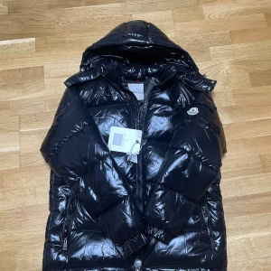 Moncler maya - Säljer en svart, glansig pufferjacka från Moncler med huva och dragkedja. Jackan har klassisk Moncler-logga på bröstet och innerfoder med serietidningsmönster. Perfekt för kalla dagar och riktigt snygg streetstil.