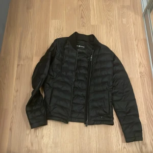 Svart pufferjacka från Sail Racing - Svart pufferjacka från Sail Racing i storlek small. Jackan har quiltad design, dragkedja framtill och diskreta loggor på insidan och vid dragkedjan. Tillverkad i syntetmaterial och har en klassisk, sportig look med lång ärm och normal passform.