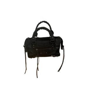 Svart handväska  - Cool svart handväska från med klassisk city-design, detaljerade nitar och dragkedja framtill. Väskan har dubbla handtag och långa dekorativa remmar på sidorna. Tillverkad i skinn med en mjuk, lite slouchy form som ger en edgy vibe.