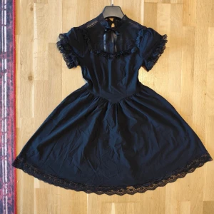 Svart kort klänning med spetsdetaljer - Supersöt svart klänning med puffiga korta ärmar och spetsdetaljer vid hals, ärmslut och nederkant. Hög hals med liten rosett och transparent parti framtill. Kjolen är utställd och har vacker spets längs fållen. Perfekt för dig som gillar romantisk och unik stil.