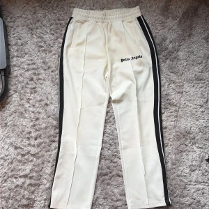 Palm Angels vita trackpants med svarta ränder Storlek L - Palm Angels vita trackpants Storlek L med svarta sidoränder och broderad logga på framsidan. Byxorna har elastisk midja, raka ben och dragkedjor vid bensluten. Tillverkade i ett glansigt syntetmaterial som ger en sportig vibe. ⚠️Dem är smutsiga med flera fläckar, därför det låga priset.
