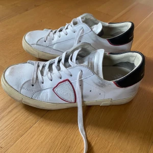 Vita Philippe Model sneakers med svarta detaljer - Snygga vita Philippe Model sneakers i skinn med svarta hälpartier och röda detaljer. Klassisk låg modell med rund tå, platt sula och snörning. Ikonisk sköldlogga på sidan och diskret branding på hälen. Perfekta för en clean och trendig look.