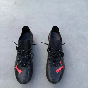 Svarta Puma Ultra fotbollsskor - Säljer ett par svarta Puma Ultra fotbollsskor med röda detaljer och logga. Skorna har snörning, låg profil och mönstrad syntetöverdel för extra grepp. Sulan har röda dobbar för bästa fäste på planen. Perfekta för dig som vill sticka ut på matchen. Skorna har aldrig använts. 