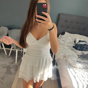 Vit omlottklänning med volanger - Supersöt vit kortklänning med omlott-v-ringning och tunna axelband. Klänningen har flera lager volanger i kjolen och är gjord i ett lätt, flowy material. Köpt från shein saknar etikett men är storlek S och är aldrig använd. Då den är för kort på mig som är 168cm.