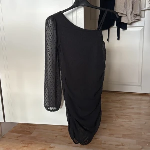 Svart oneshoulder klänning Gina Tricot - Svart oneshoulder klänning från Gina Tricot med lång ärm i transparent prickigt tyg och rynkade detaljer längs sidorna. Klänningen har en smal axelrem och är figurnära med snygg struktur i materialet. Perfekt för dig som vill sticka ut med en cool look.