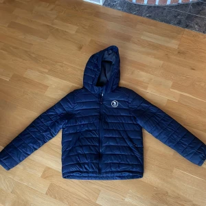 Mörkblå pufferjacka Abercrombie Kids - Snygg mörkblå pufferjacka från Abercrombie Kids med huva och dragkedja. Jackan har ett broderat vitt logomärke på bröstet och är quiltad för extra värme. Perfekt för kyliga dagar och har en stilren look som funkar till det mesta. En mindre slitning på bröstet men inget som syns vid andvändning. 