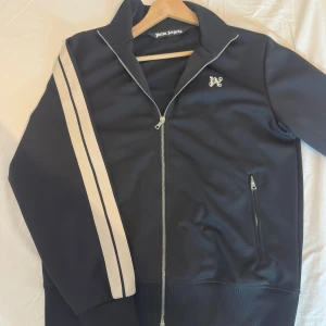Svart track jacket från Palm Angels - Svart track jacket från Palm Angels med vita dubbla ränder längs ärmarna, hel dragkedja framtill och två fickor med dragkedja. Jackan har en sportig look, hög krage och logga på bröstet. Perfekt för en streetwear-stil. Nypris 5500kr