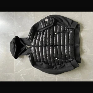 Svart pufferjacka med huva från Moncler - Svart pufferjacka från Moncler med quiltad framsida, huva med snörning och dragkedjor på fickorna. Jackan har ribbade muddar och en patch med Moncler-logga på ärmen. Materialmix av polyester och bomull ger både värme och stil. Passar M fast lite stor kanske 