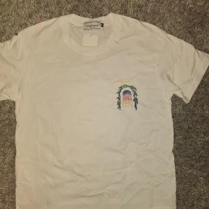 Vit t-shirt från Casablanca med tryck - Vit t-shirt från Casablanca med färgglatt tryck av en byggnad och gröna blad på bröstet och ryggen. T-shirten har rund hals, korta ärmar och är gjord i mjuk bomull. Perfekt för dig som gillar stilrena plagg med unik design.