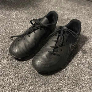 Svarta Nike Phantom fotbollsskor  - Säljer ett par svarta Nike Phantom fotbollsskor med klassisk Swoosh-logga i vitt på sidan. Skorna har snörning, rund tå och platt sula med fasta dobbar för bra grepp på planen. Ovandelen är i syntetmaterial med diskreta mönster för extra bollkontroll.