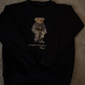 Svart sweatshirt Polo Ralph Lauren Bear - Svart sweatshirt från Polo Ralph Lauren med ikoniska björnmotivet på bröstet. Tröjan har rund halsringning, långa ärmar och är tillverkad i 100% bomull. Perfekt för dig som gillar streetstyle och klassiska designer.