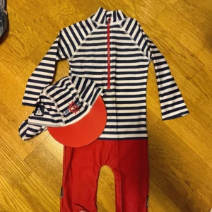 Randig keps med röd skärm - Badkläder barn -74 - Snygg keps med marinblå och vita ränder samt röd skärm. Kepsen har ett broderat motiv med kompass och segelbåt på sidan. Perfekt för soliga dagar vid stranden eller poolen. Uv kläder barn unisex str 74 cm 9-12 månader 