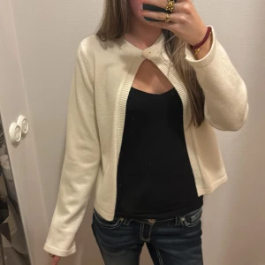 Krämvit kofta med knapp - Säljer en krämvit kofta med lång ärm och en stor dekorativ knapp vid halsen. Koftan har öppen front och ribbade kanter, perfekt att slänga över ett linne eller t-shirt. Enkel och stilren design som passar till jeans eller kjol.