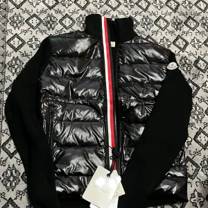 Svart dunväst från Moncler - Svart dunväst från Moncler med glansig quiltad framsida och stickade svarta sidor. Västen har en dragkedja med röd och vit rand längs öppningen samt Moncler-logga på axeln. Materialet är dun och polyester, perfekt för kyliga dagar.