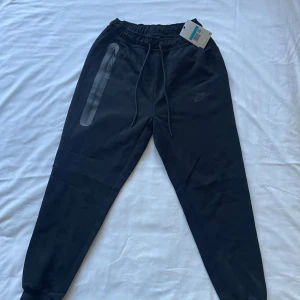 Nike tech byxor svart - Svarta tech mjukisbyxor från Nike i storlek M. Byxorna har dragsko i midjan, ribbade muddar vid bensluten och en snygg dragkedjeficka på ena benet. Perfekta för chill eller träning, med diskret Nike-logga på låret.