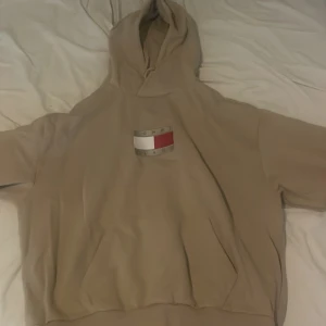 Äkta Beige hoodie från Tommy Jeans - Säljer en beige hoodie från Tommy Jeans med klassisk logga på bröstet. Tröjan har huva, känguruficka och är tillverkad i mjuk bomull. Perfekt för dig som gillar en clean och stilren look. Storlek M/L