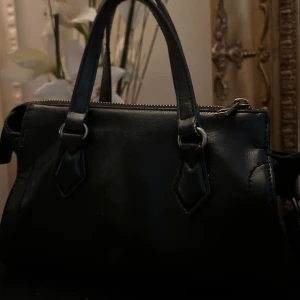 Svart handväska i skinn med dragkedja - Zara - Snygg svart liten handväska i skinn med stilrena detaljer i silverfärgad metall. Väskan har två handtag och en avtagbar axelrem, samt dragkedja upptill. Perfekt storlek för det viktigaste och passar till många olika looks. HELT SLUTSÅLDA & Inhandlades för 450 kr, men jag säljer för billigare!💕