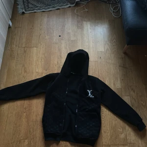 Svart Louis Vuitton hoodie med monogram - Svart hoodie från Louis Vuitton med klassiskt LV-monogram i tyget och broderad logga på bröstet. Tröjan har dragkedja framtill, två stora fickor och huva. Perfekt för dig som vill ha en exklusiv streetstyle-look. 