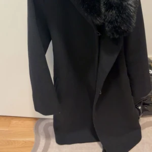 Svart kappa med pälsdetalj - Snygg svart kappa med stor krage i pälsimitation. Klassisk rak modell med långa ärmar och diskreta fickor på sidorna. Perfekt för dig som vill ha en stilren och elegant look under höst och vinter.