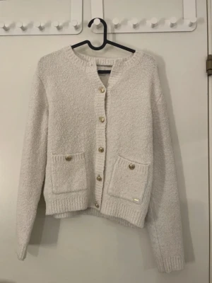 Vit kofta från Holly & Whyte - Mysig vit kofta från Holly & Whyte med guldfärgade knappar och två fickor framtill. Koftan är stickad i ett fluffigt material och har ribbade muddar vid ärmslut. Orginal pris är 500kr