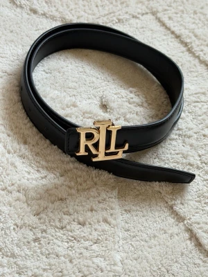 Svart skinnbälte från Ralph Lauren - Snyggt svart bälte från Ralph Lauren med ett stilrent RL-spänne i guld. Bältet är gjort i slätt skinn och har flera hål för justerbar passform. Perfekt accessoar för att lyfta din outfit med en lyxig touch.