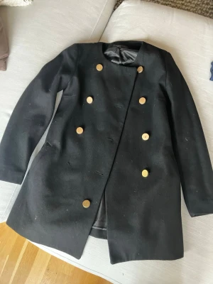 Svart ullblandad skepparkavaj med guldknappar - Snygg svart skepparkavaj med dubbelknäppning och stora guldiga knappar framtill. Jackan har en rak och klassisk siluett, helfodrad insida och långa ärmar. Perfekt för dig som vill ha en stilren look med lite edge. En knapp har lossnat men kan enkelt sys på i övrigt fint skick. 61 % ull. 