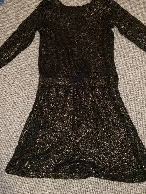 Glittrig svart och guld klänning Name It - Supersnygg svart klänning från Name It med guldglittrande detaljer. Klänningen har lång ärm, markerad midja med snörning och dragkedja i ryggen. Perfekt för dig som vill sticka ut lite extra!