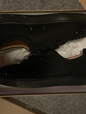  Christian Louboutin - Säljer ett par svarta sneakers från Christian Louboutin med ikonisk röd sula och svarta nitar på tån. Skorna har snörning och är tillverkade i mocka med insida i beige skinn. Perfekt för dig som gillar edgy och lyxig stil.