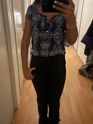 Blå mönstrad blus med knappar - Säljer en blå blus med vitt paisleymönster och korta ärmar. Toppen har knappar framtill och en liten rosett i rött vid knappraden. Materialet känns lätt och luftigt, perfekt för varmare dagar. Märke syns på etiketten nertill.