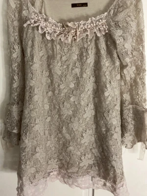 Beige spetsklänning från Soma - Supersöt beige kortklänning från Soma med spets ärmar och blommigt spetsmönster. Klänningen har volangdetaljer vid ärmslut och nederkant samt en fin rosett och spets vid halsringningen. Perfekt för dig som gillar romantisk stil och detaljer.