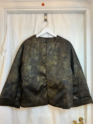 Svart vadderad jacka i 100% mullbärssilke - Svart vadderad jacka i 100% mullbärssilke  Elegant svart jacka med diskret mönster. Yttertyget är gjort av 100% mullbärssilke, med en vacker, lågmäld struktur som syns olika beroende på ljuset.  Jackan är lätt vadderad med quiltat foder, vilket gör den varm men inte klumpig – perfekt för höst och vinter. Modellen har nedhasad axel (loose fit) som ger en avslappnad och bekväm känsla samt bra passform för olika kroppstyper.  Dekorativa guldfärgade knappar framtill ger jackan en subtil, elegant det