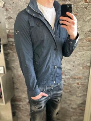 Blå vindjacka från J.Lindeberg - Snygg marinblå vindjacka från J.Lindeberg med krage och dragkedja samt knappar framtill. Jackan har flera fickor med lock och knappdetaljer, samt ett diskret emblem på ärmen. Perfekt för dig som gillar stilren och sportig look. OBS! Inte mina bilder utan dem är från Bondeclothing