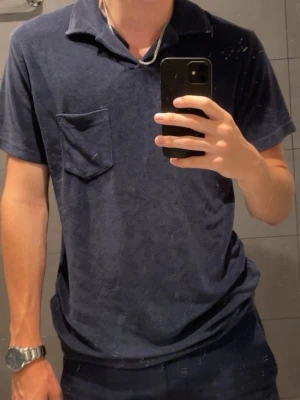 Mörkblå frotté polotopp - Säljer en mörkblå polotopp i mjuk frotté med korta ärmar och en bröstficka. Toppen har en klassisk krage och avslappnad passform, perfekt för en chill och stilren look. Passar dig som gillar enkel men snygg design.